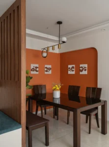 Dining area with a bold burnt orange arched colour-block accent wall, four abstract art prints, a dark wood dining table with six chairs, and a sculptural pendant light with brass cylinders and globe shades.