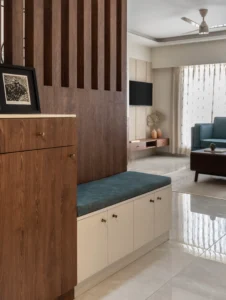 Apartment foyer with a dark walnut vertical slatted partition wall, a wooden cabinet with brass knobs, and a built-in storage bench with a teal upholstered cushion, opening into the living room beyond.