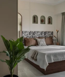 Wide-angle view of a guest bedroom with sage green arched wall niches housing carved wooden artefacts above the bed, a patterned fabric headboard, grey bedding, sage green sheer curtains, and a lush indoor plant in the foreground.