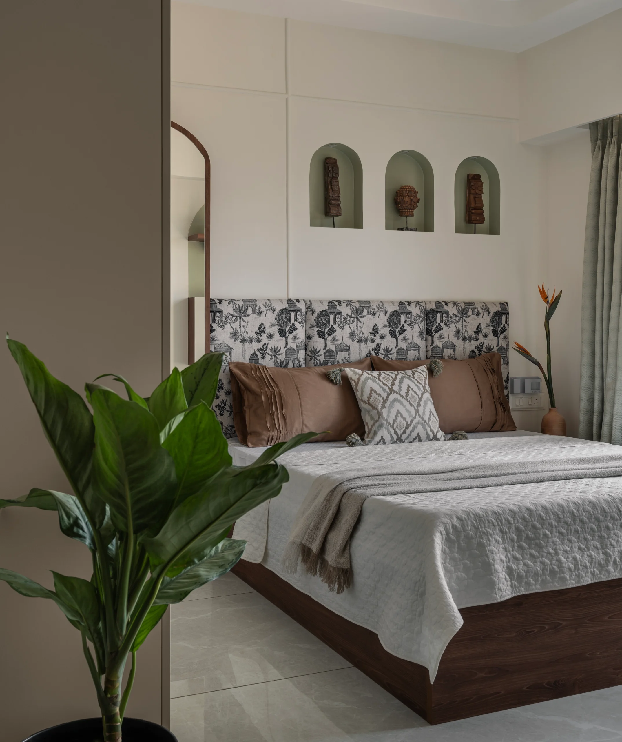 Wide-angle view of a guest bedroom with sage green arched wall niches housing carved wooden artefacts above the bed, a patterned fabric headboard, grey bedding, sage green sheer curtains, and a lush indoor plant in the foreground.