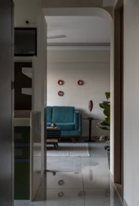 View through a rounded arched doorway into a sitting area with a teal velvet armchair, a carved African mask on a tall black pedestal table, a fiddle leaf fig plant, and terracotta wall plates.