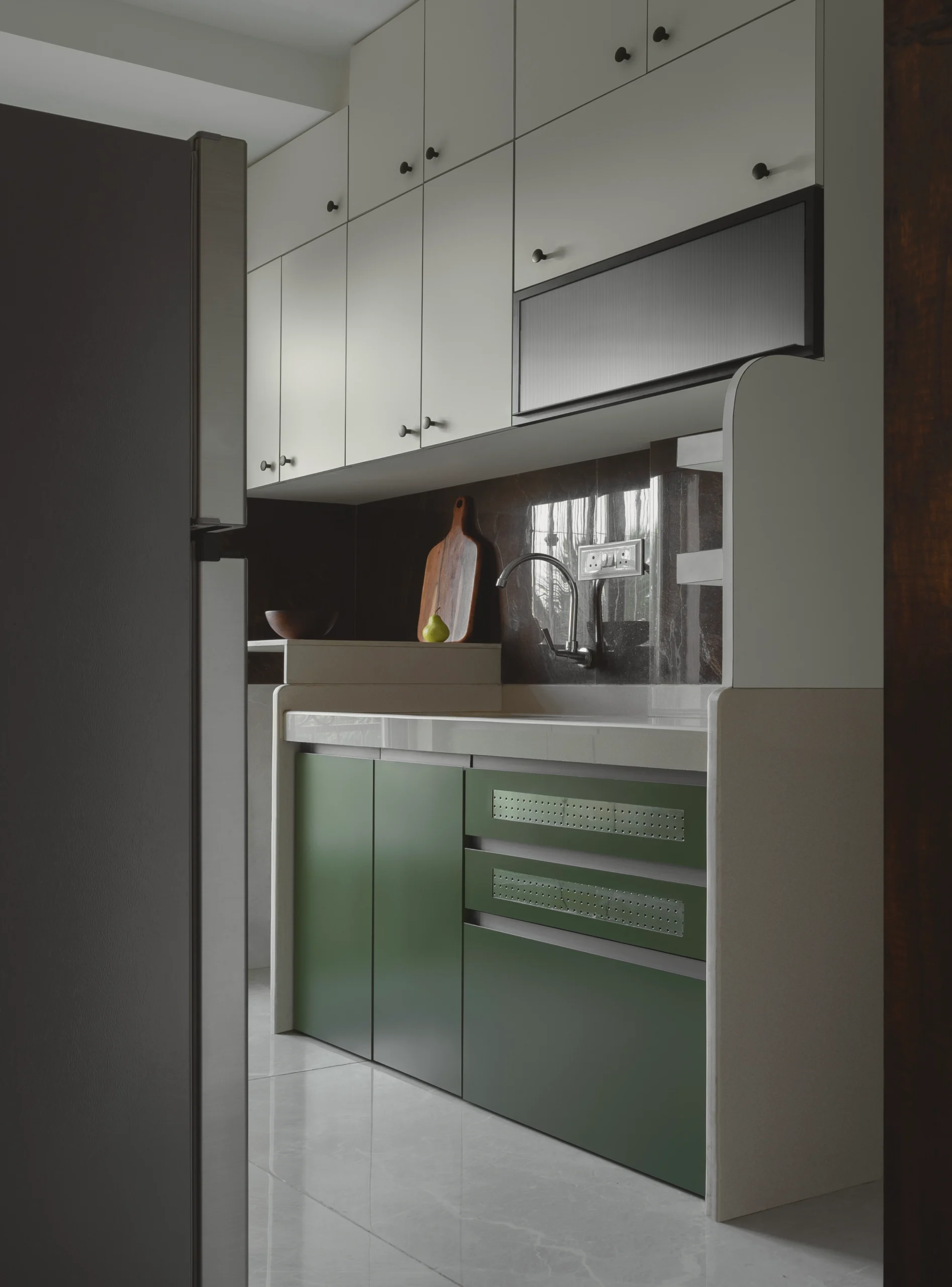 Two-tone modular kitchen with cream upper cabinets, sage green lower cabinets with perforated drawer fronts, a dark stone backsplash, and a chrome gooseneck faucet in a contemporary Indian apartment.