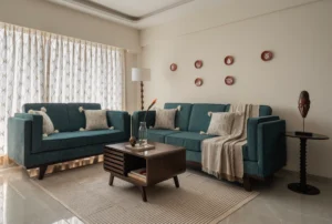 Contemporary Indian living room with teal velvet sofas, a dark walnut mid-century style coffee table, a natural fibre rug, terracotta decorative plates on the wall, and sheer geometric-print curtains.