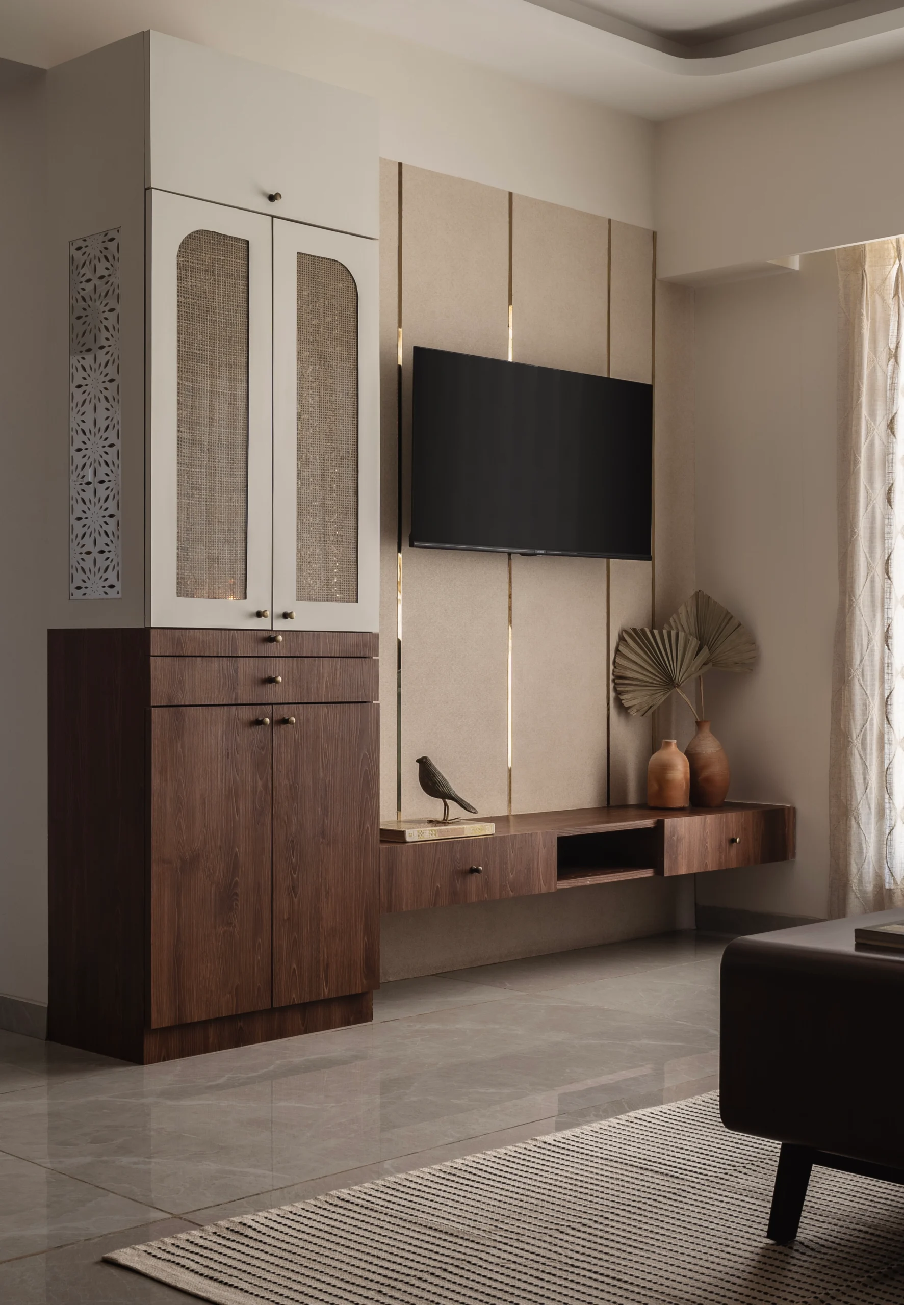 Living room TV wall featuring a warm beige textured panel with vertical brass strips, a floating walnut media console with terracotta vases, and a full-height white storage cabinet with arched cane-panelled doors.
