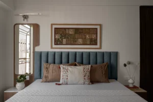 Master bedroom with a steel-blue channel-tufted upholstered headboard, brown and white decorative cushions, and a framed Kantha patchwork textile artwork mounted on the wall above the bed.