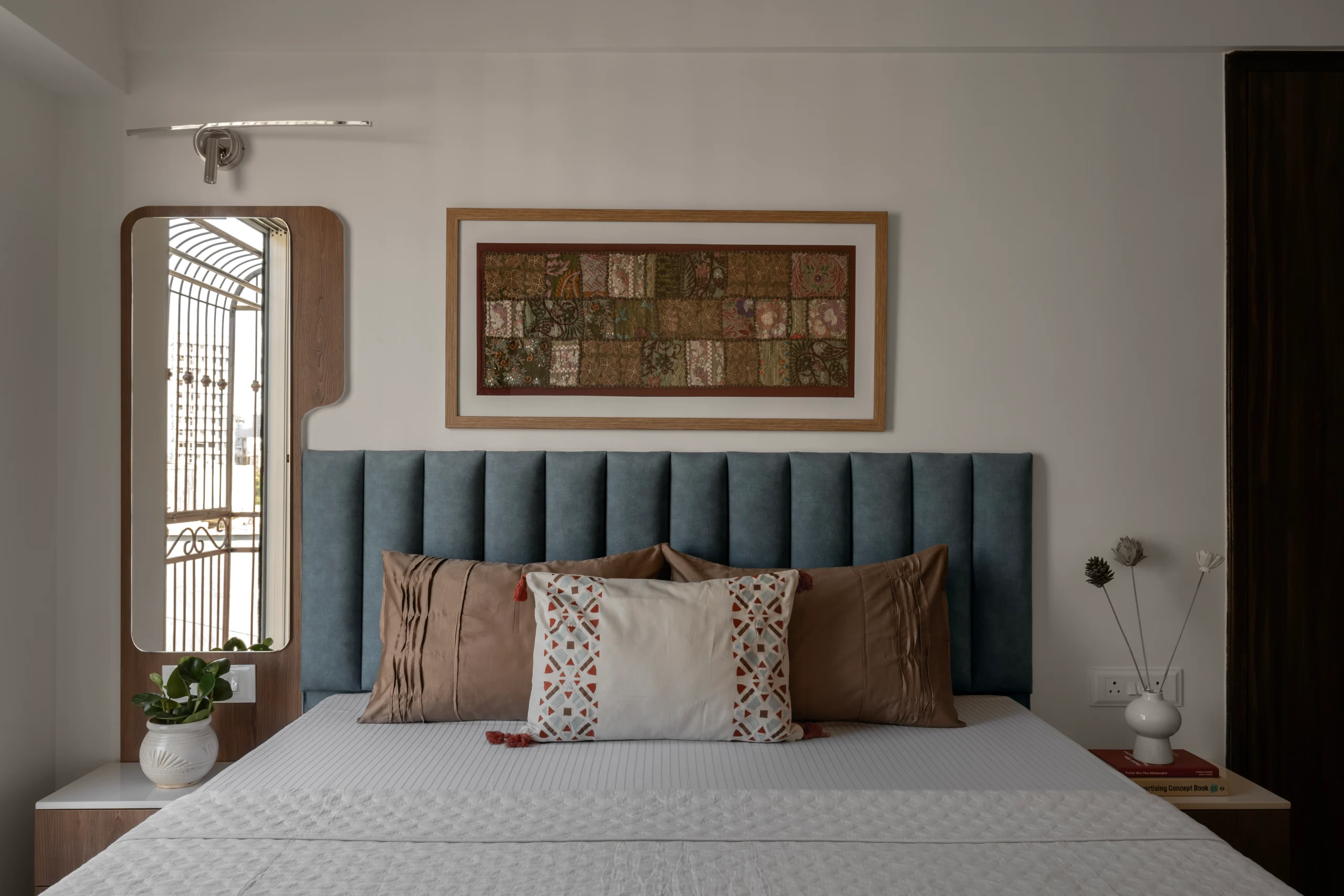 Master bedroom with a steel-blue channel-tufted upholstered headboard, brown and white decorative cushions, and a framed Kantha patchwork textile artwork mounted on the wall above the bed.