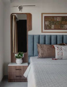 Close-up of a master bedroom nightstand in walnut wood with a white countertop, a green indoor plant in a ceramic pot, and a tall full-length arched mirror set in a walnut wood frame beside the bed.