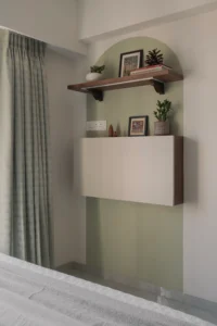 Wall-mounted fold-down desk in closed position against a sage green arched accent wall, with a decorative wooden shelf above styled with books, a pinecone, and a small white plant in a Mumbai apartment bedroom.