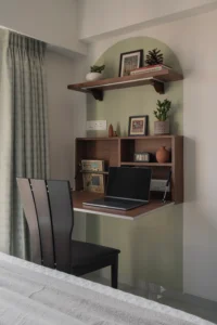 Open wall-mounted fold-down workstation in a bedroom with a laptop, walnut wood storage cubbies, a terracotta vase, and a task lamp, transforming a compact corner into a functional home office.